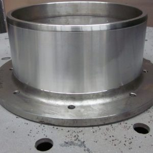 A large metallic cylinder with a wide flange sits on a flat, round metal surface that has evenly spaced small holes. The flange has several holes around its edge and some scattered metal debris nearby.