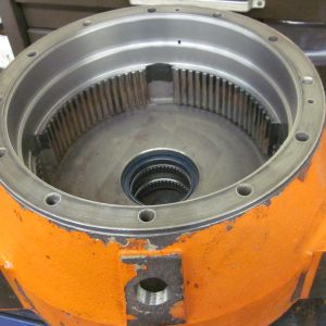 A close-up of a large orange metal mechanical part, possibly a gear housing or transmission casing, with visible ball bearings and cylindrical rollers inside, resting on a workbench with tools and parts nearby.