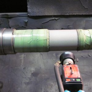 A cylindrical metal shaft clamped in a machine, partially wrapped with green tape, with a red measurement instrument and cables positioned in front of it. The background is industrial and metallic.