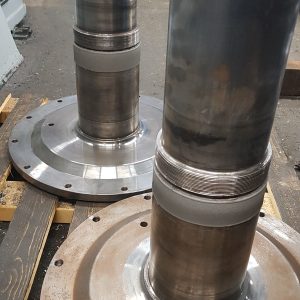 Two large, cylindrical metal shafts with flanges are placed vertically on a wooden pallet. Each shaft has a circular base with bolt holes and an eye bolt on top. The background includes a blue wall and machinery.