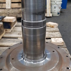 A large metal shaft with a wide, circular base sits upright on a wooden pallet. The shaft appears to be part of industrial machinery, with visible machining marks and a few bolts around the base.