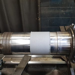 A large metal shaft mounted on a lathe, with a gear on one end and a textured section in the middle. A gauge and metal tools are visible nearby, and the background is an industrial workshop.