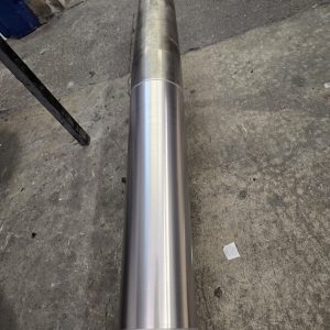 A large, cylindrical metal shaft is lying horizontally on a concrete floor. The shaft has a polished, shiny section in the center, contrasting with the duller, worn ends.
