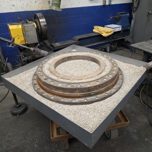 A large metal ring with bolts is placed on a square tray filled with gravel in a workshop. Tools, machines, and workbenches are visible in the background.