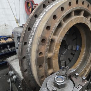 A large metal flange with multiple bolt holes is mounted on a lathe in a workshop, showing signs of wear and machining. Tools and equipment are visible in the background.