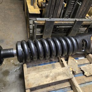 A large, heavy-duty black metal coil spring assembly resting on a wooden pallet in an industrial workshop, with forklift parts and tools visible in the background.