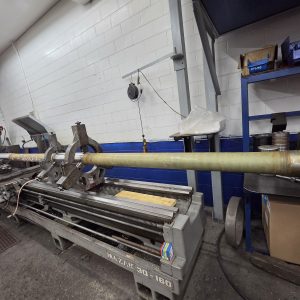 A long cylindrical object is mounted on a large metal lathe in a workshop with white and blue walls, shelves with tools, and fluorescent lights overhead.