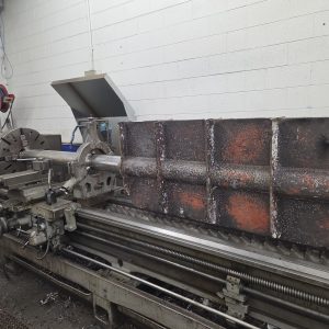 A large metal shaft with a rusted, rectangular paddle is mounted on an industrial metal lathe in a workshop with white brick walls.