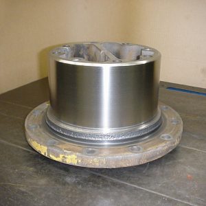 A cylindrical metal machine part with a shiny surface and a wide, round, flat base sits on a metal table. The base has multiple evenly spaced bolt holes around its edge. The background is plain.