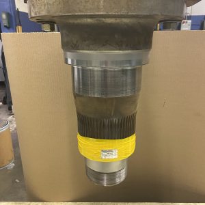 A large metal shaft with a flanged top is positioned vertically in front of a cardboard background. The lower part of the shaft is partially wrapped with yellow tape. Industrial equipment is visible in the background.