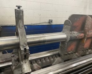 A metalworking lathe machine is shaping a cylindrical metal rod, with metal shavings visible around the tool, inside an industrial workshop with white and blue walls.
