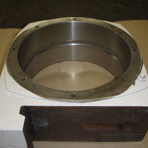 A large metal ring with bolt holes sits partially embedded in white foam packaging on a cardboard surface, with a wooden or metal bracket attached to one side.