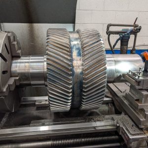 A large metal gear with helical teeth is mounted on a lathe machine in an industrial workshop, ready for machining. The background shows a white brick wall and various mechanical components.