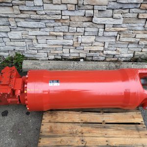 A large red hydraulic cylinder lies horizontally on a wooden pallet outdoors, with a stone wall in the background.