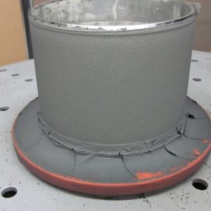 A cylindrical metal object with a gray, matte-coated surface stands on a perforated metal table. The base has uneven, cracked coating with traces of orange underneath.