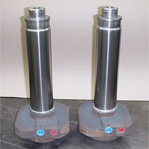 Two shiny metal hydraulic cylinders with cylindrical shafts and rusted bases, each with a red and a blue fitting, standing upright on a dark surface against a plain light background.